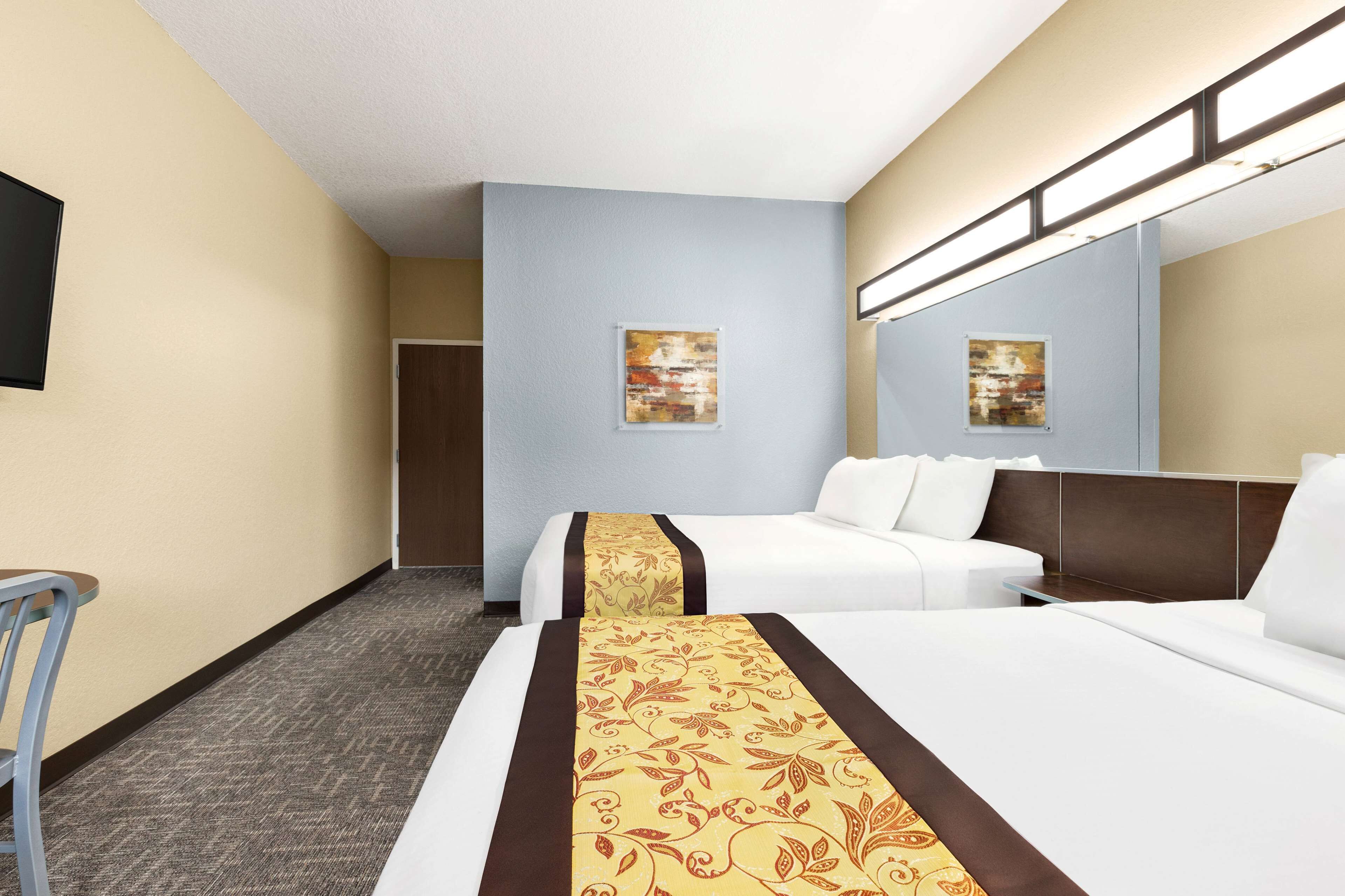 Quarto de hotel com duas camas, uma secretária e uma cadeira. Há duas pinturas nas paredes e um espelho grande.