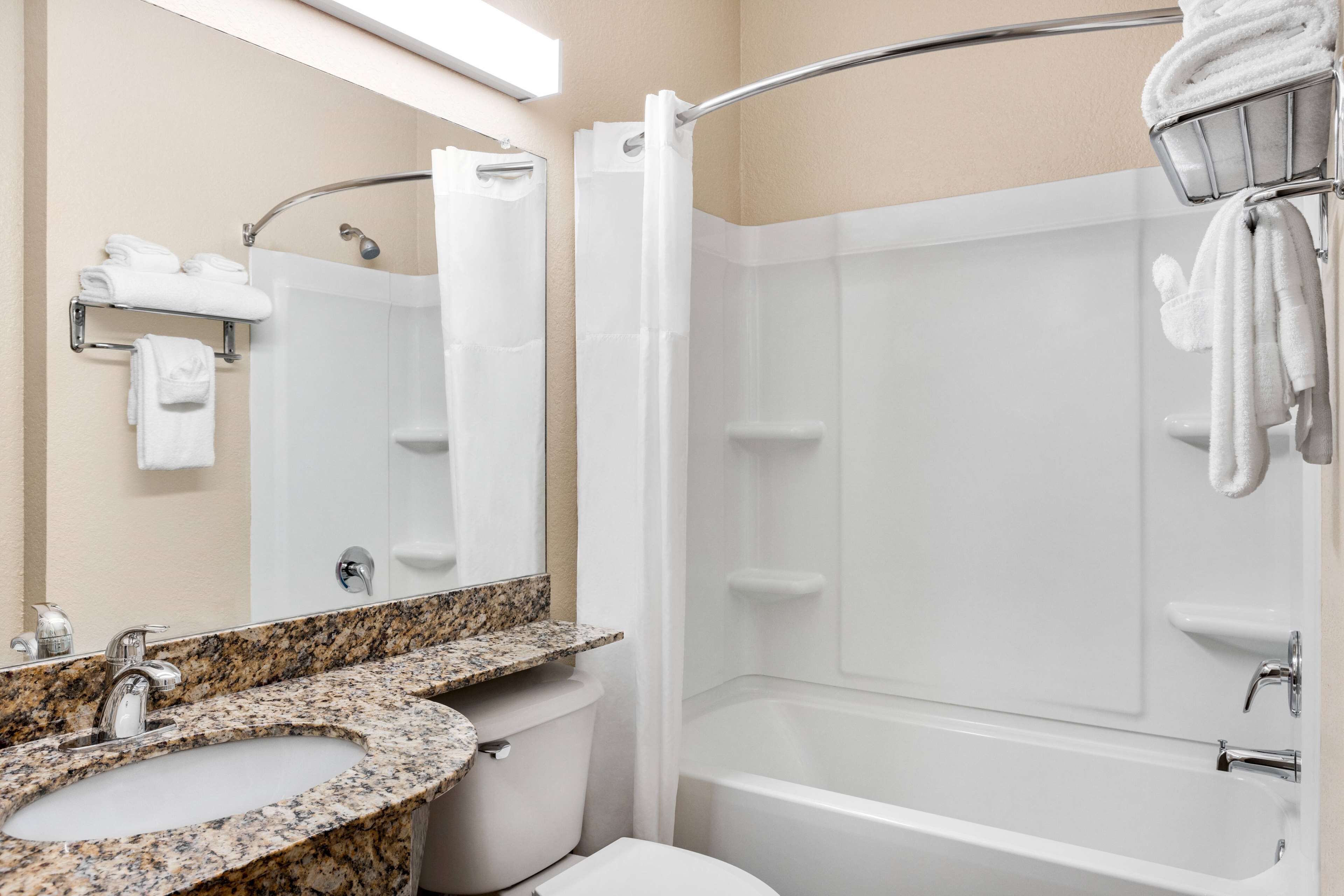 Une salle de bain moderne avec un lavabo surmonté d’un plan de travail en granit, des toilettes blanches, une baignoire et un rideau de douche.