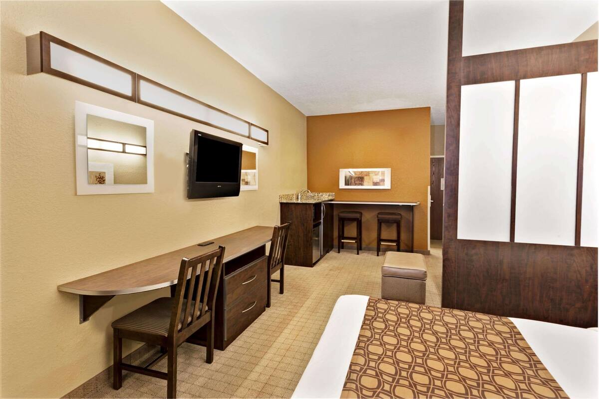 Une chambre d’hôtel compacte comprenant un coin cuisine, un téléviseur à écran plat, un bureau avec une chaise et un lit recouvert d’une courtepointe à motifs.