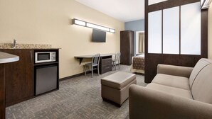 Een compacte hotelkamer met een kitchenette, een flatscreen televisie, een bureau met stoelen, een bank en een groot raam.