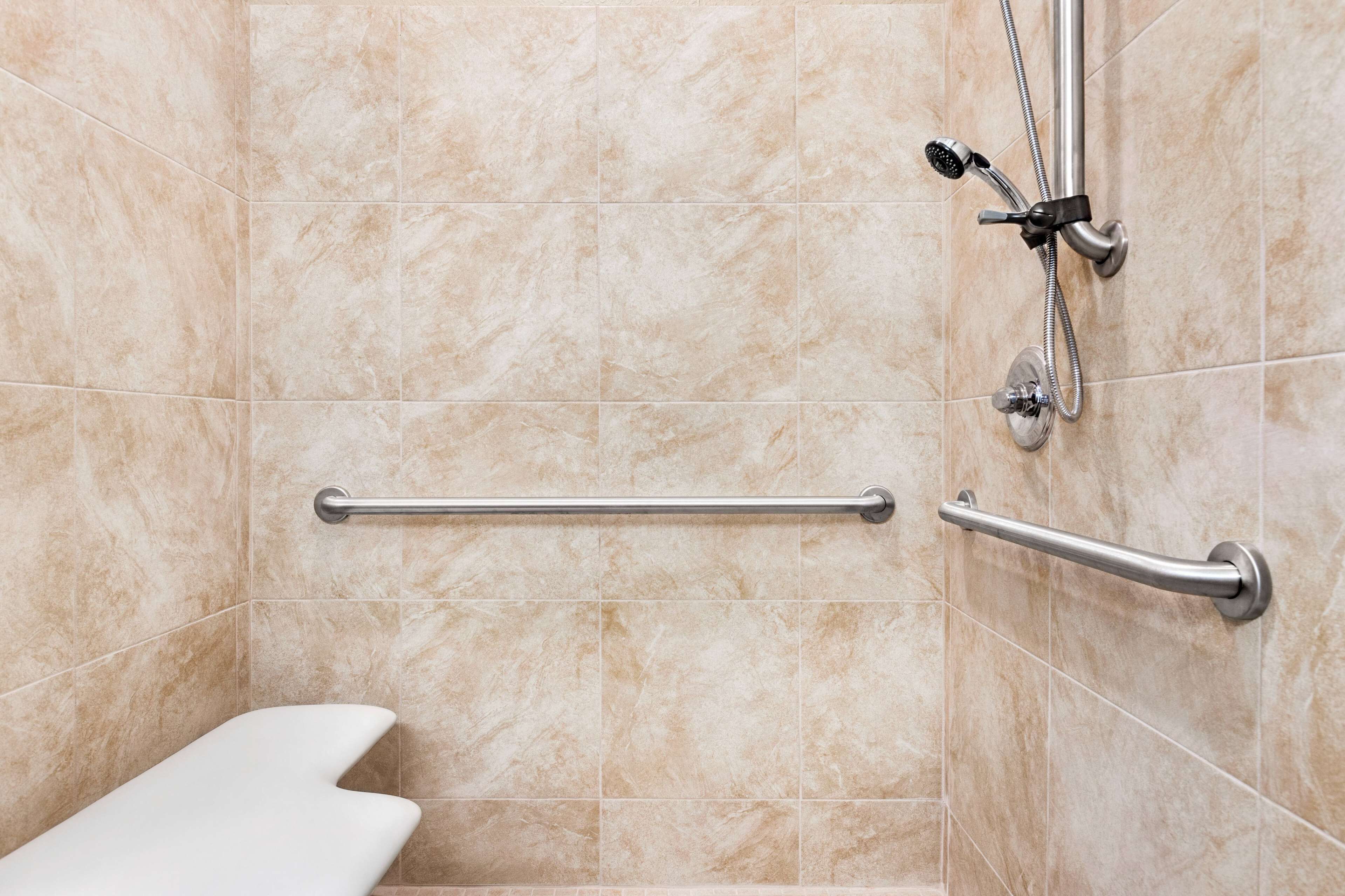 A bathroom with beige tiles, a white bathtub, and a shower with a handheld showerhead and grab bars.