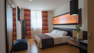 A hotel room with a bed, bedside table, a suitcase, a mirror, a TV, and a window with curtains.