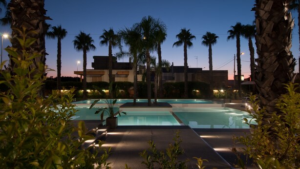 A swimming pool area at dusk with palm trees and a building in the background. - Sanlu Hotel (Carpignano Salentino)