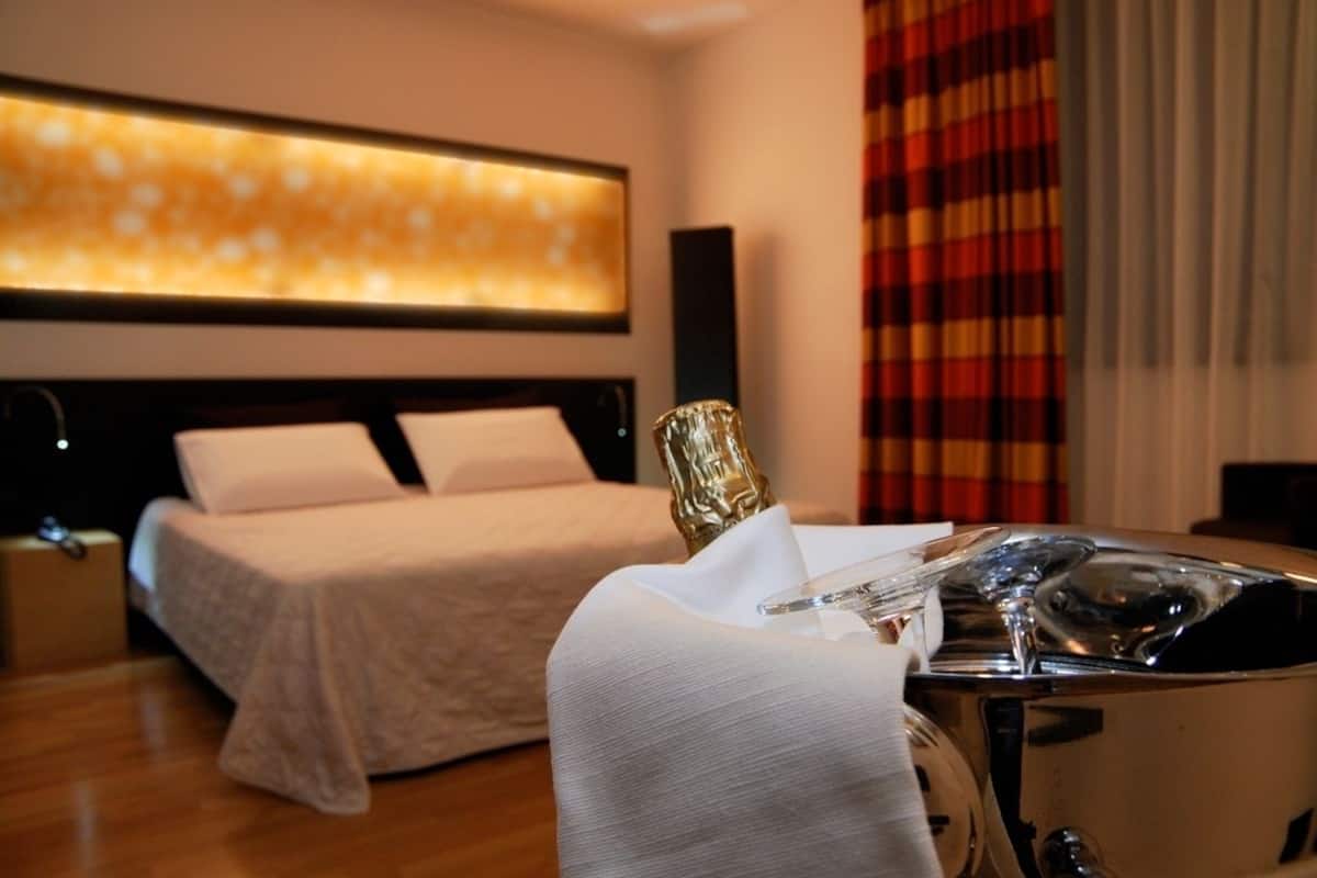 Quarto de hotel com uma cama, uma mesinha de cabeceira com um frasco e um candeeiro fixado na parede.