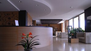 A modern reception area with a curved desk, potted plants, and a seating area with a sofa and armchairs. - Sanlu Hotel (Carpignano Salentino)