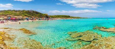 A beach with clear turquoise water, rocky formations, and a crowd of people.