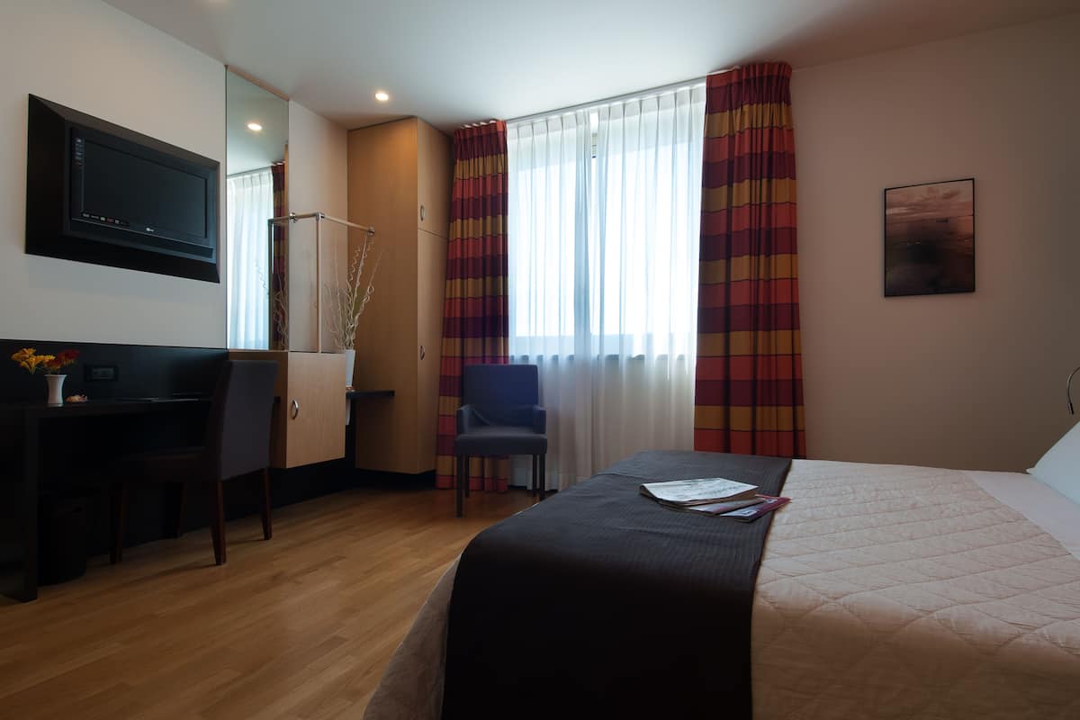 Quarto de hotel com cama, escrivaninha, cadeira, televisão e janela com cortinas.