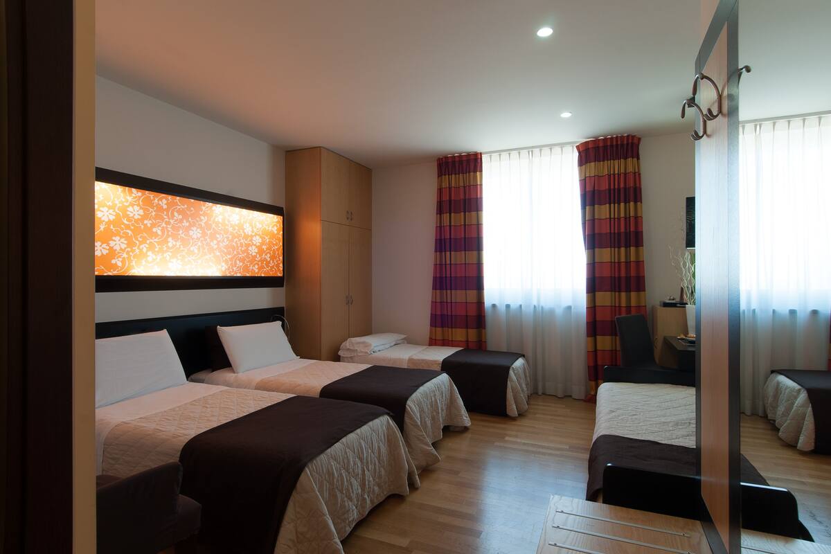 Quarto de hotel com duas camas, uma escrivaninha, uma cadeira e uma janela grande com cortinas.