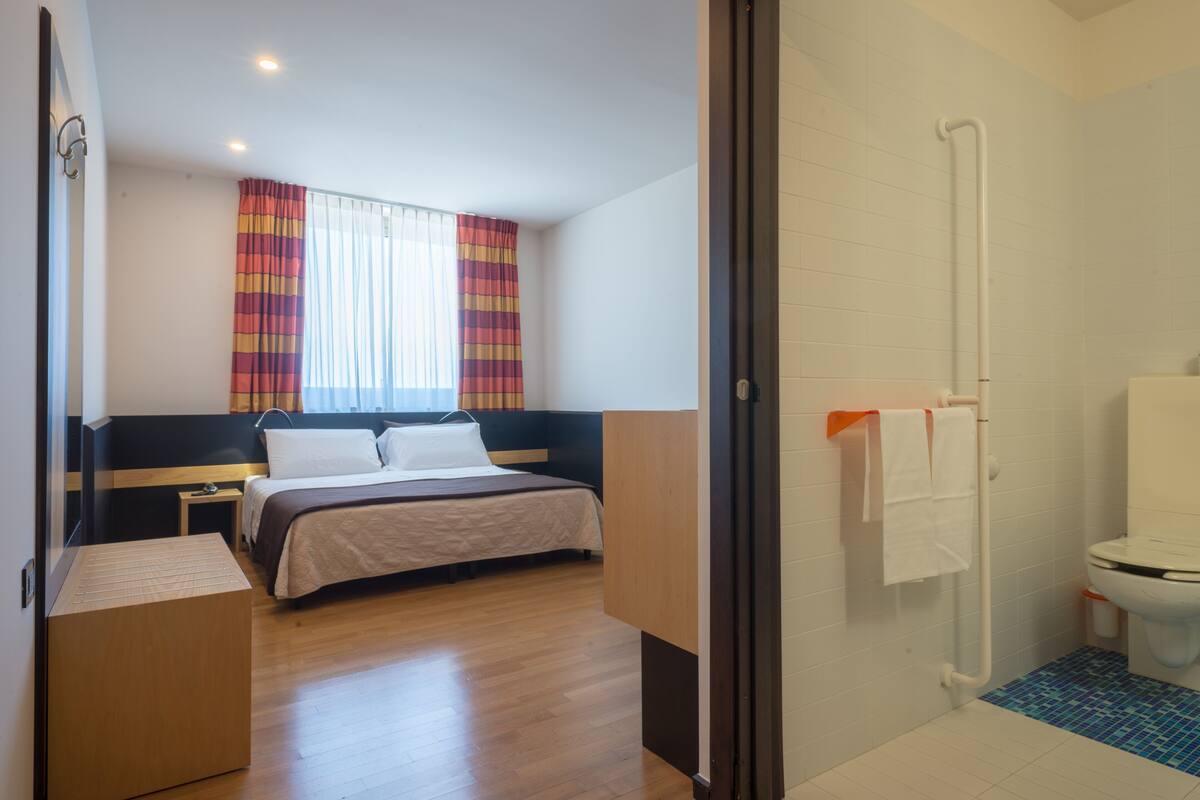 Quarto de hotel com cama, mesa de cabeceira, janela com cortinas e banheiro com vaso sanitário e chuveiro.