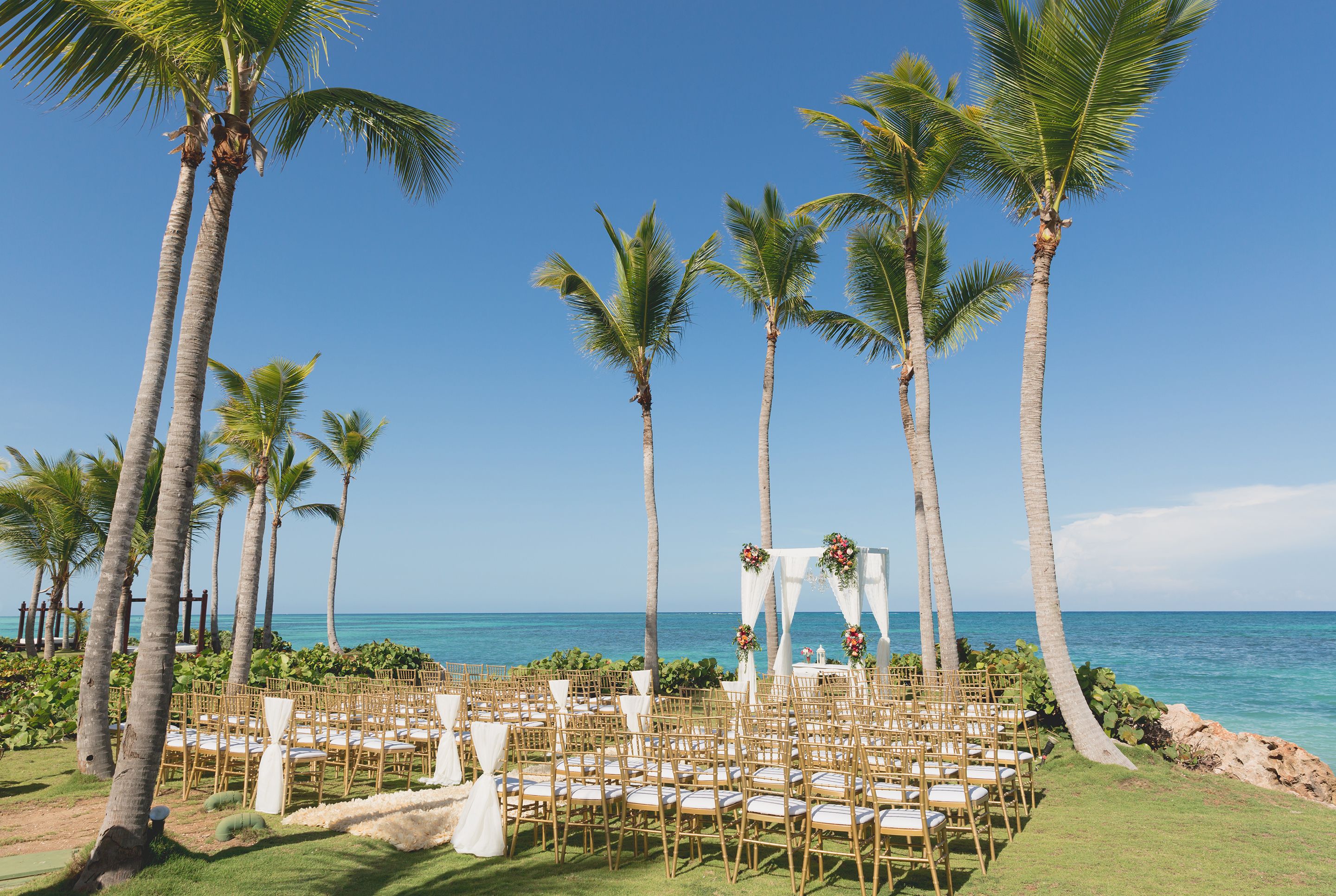 A beach wedding setup with a circular arrangement of chairs and a white arch adorned with flowers, palm trees, and a view of the ocean.