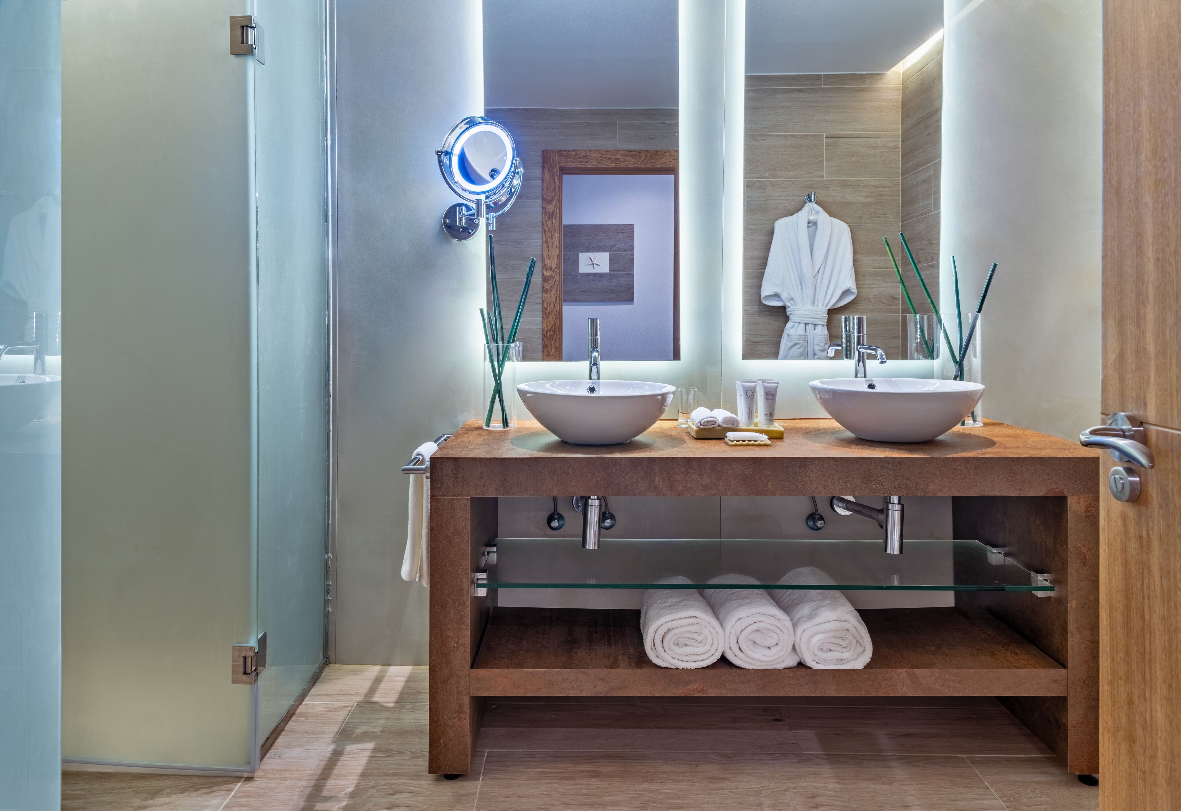 A modern bathroom with two sinks, a glass shelf with towels, and a mirror with a light.