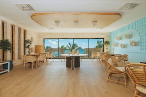 A spacious room with a large round table and wicker chairs, large windows with a view of palm trees, and a ceiling with a unique design.