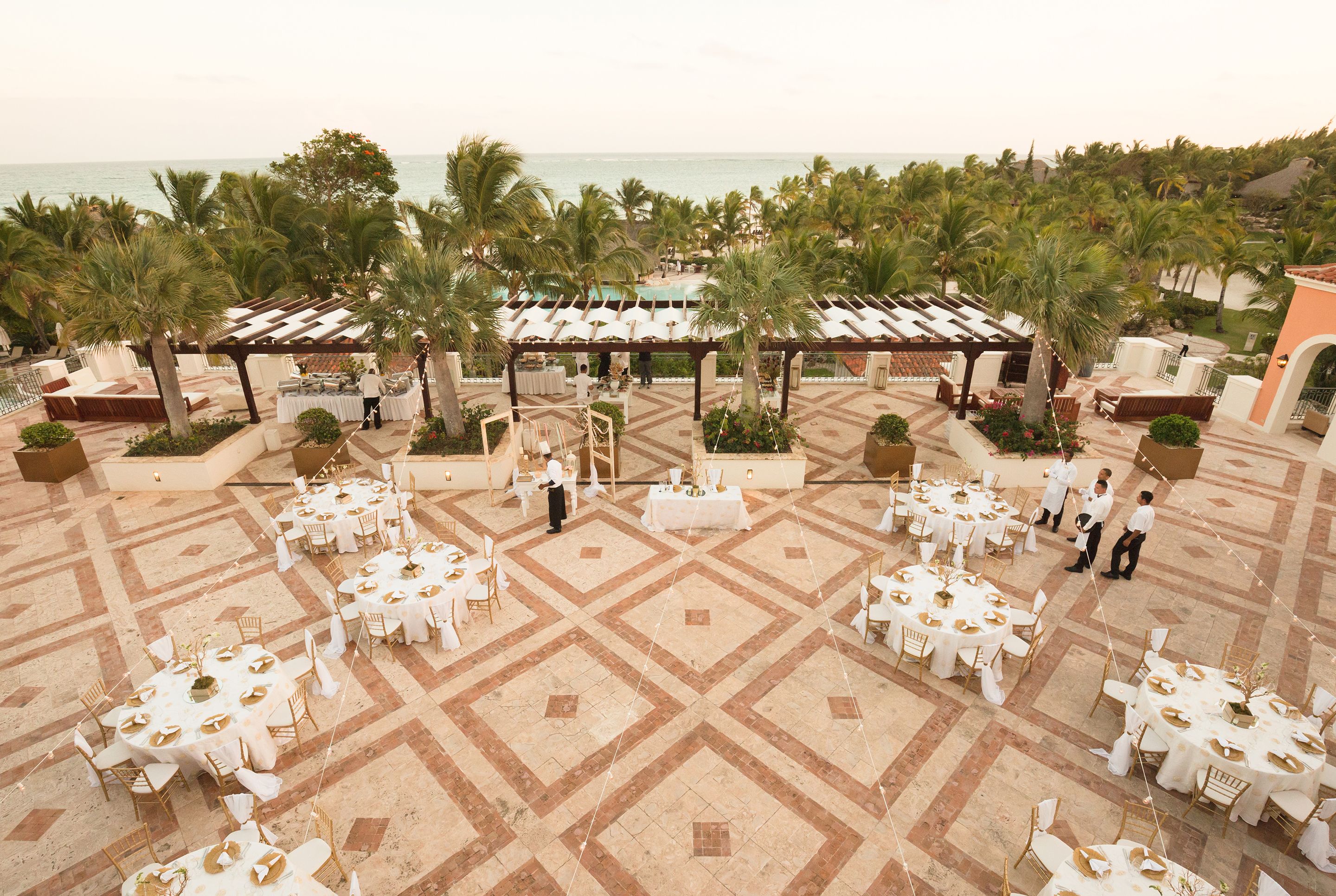 An outdoor dining area with multiple round tables set up for guests, surrounded by palm trees and a view of the ocean.
