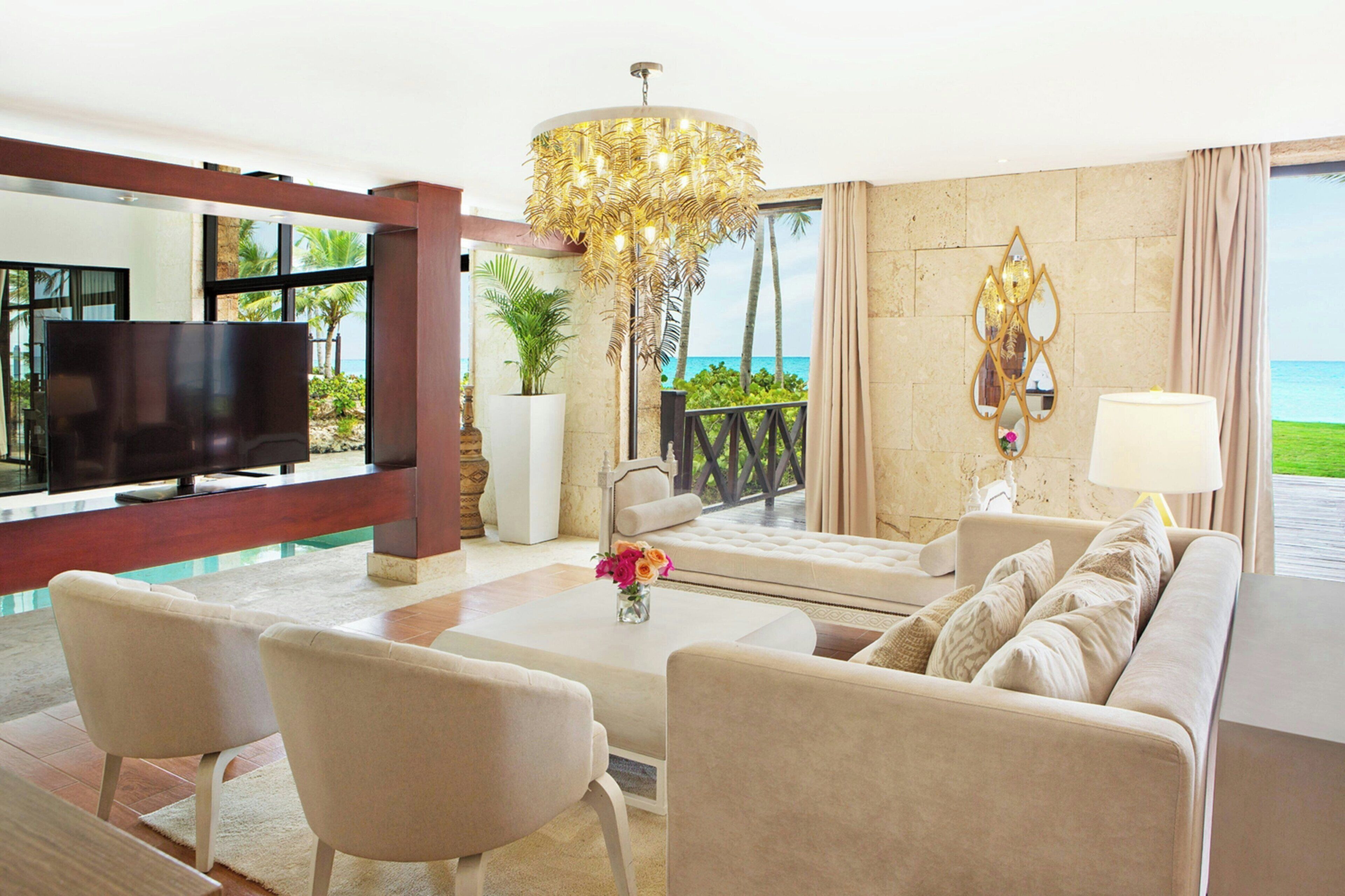 A modern living room with a large flat-screen TV, comfortable seating, a decorative chandelier, and a view of the ocean.