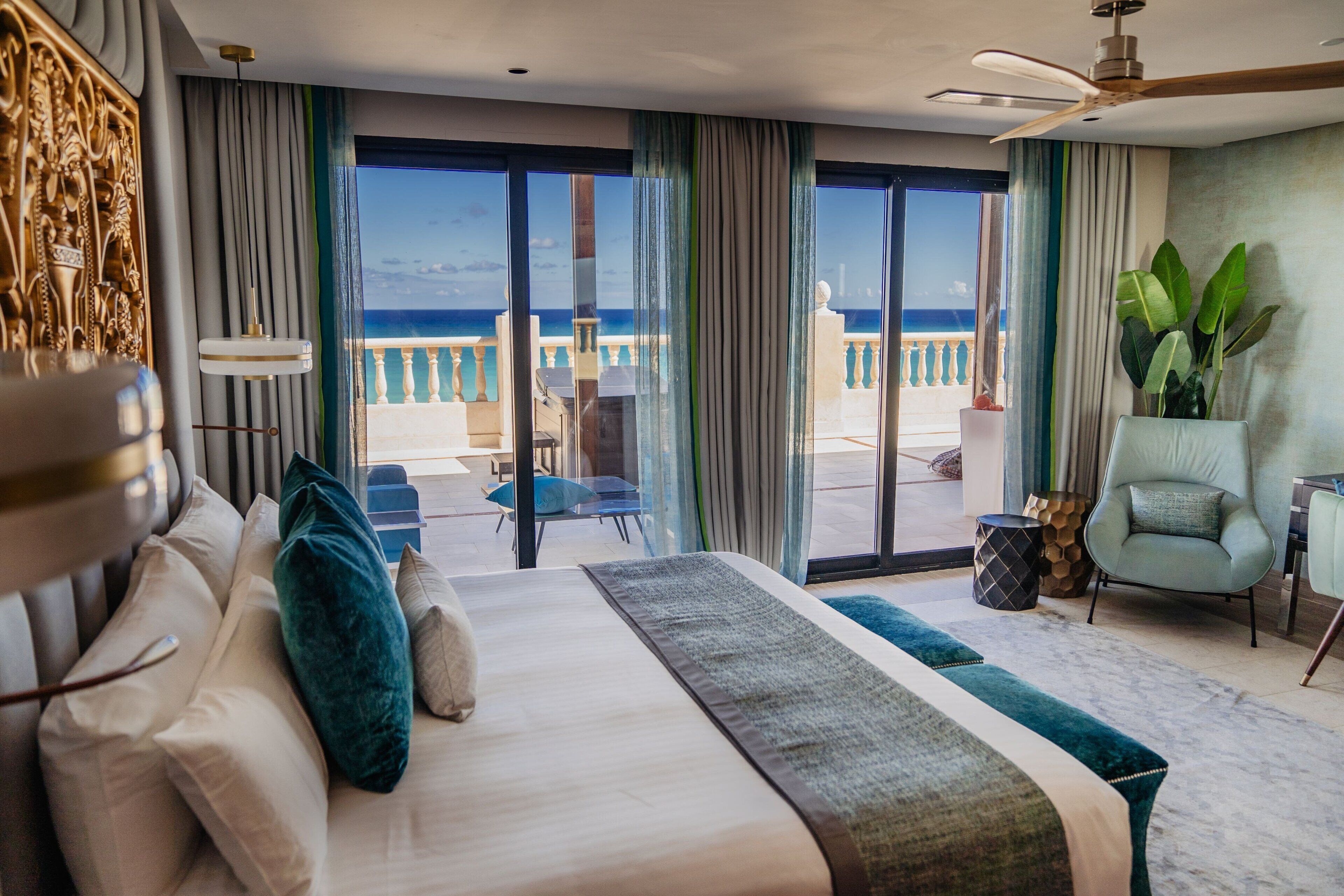 A hotel room with a large bed, a balcony with a view of the sea, a ceiling fan, and a chair.