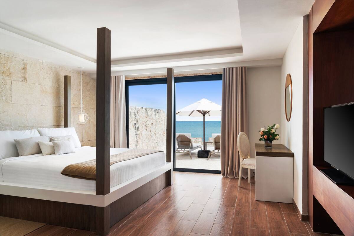 A modern hotel room with a large bed, a desk, and a TV. The room has a view of the sea and a balcony with outdoor seating.
