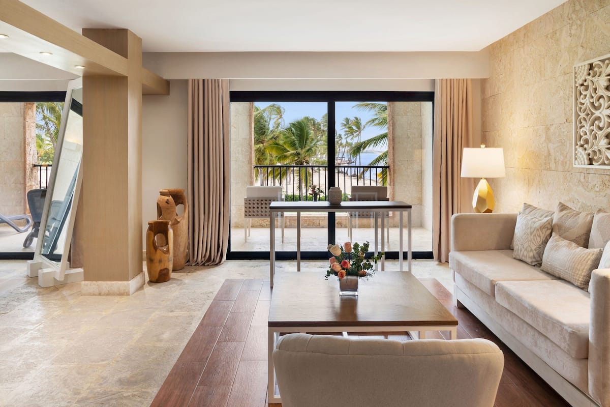 A modern hotel room with a balcony, a sofa, a coffee table with flowers, and a view of palm trees and the sea.