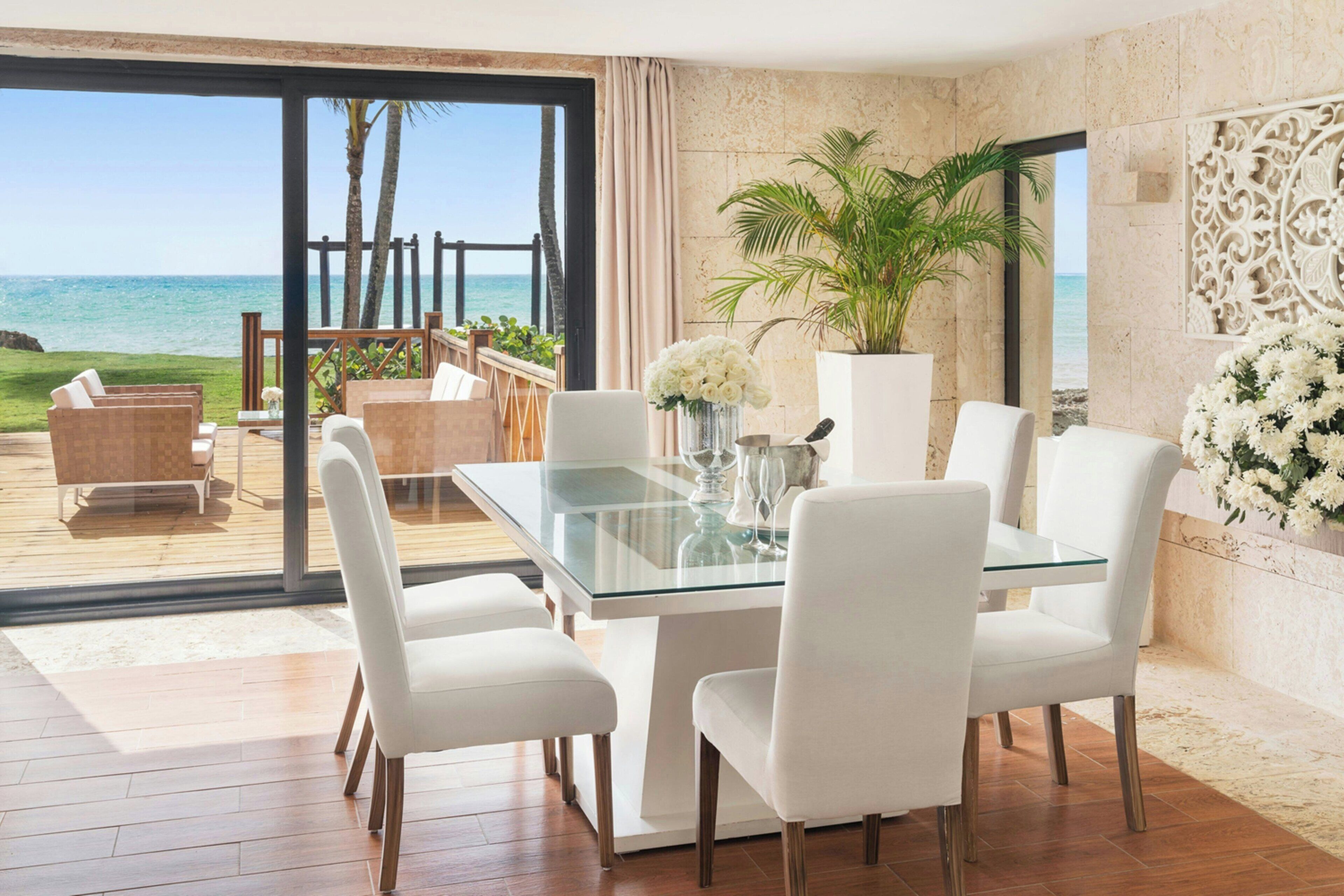 A modern dining area with a glass table and white chairs, overlooking a beach with palm trees.