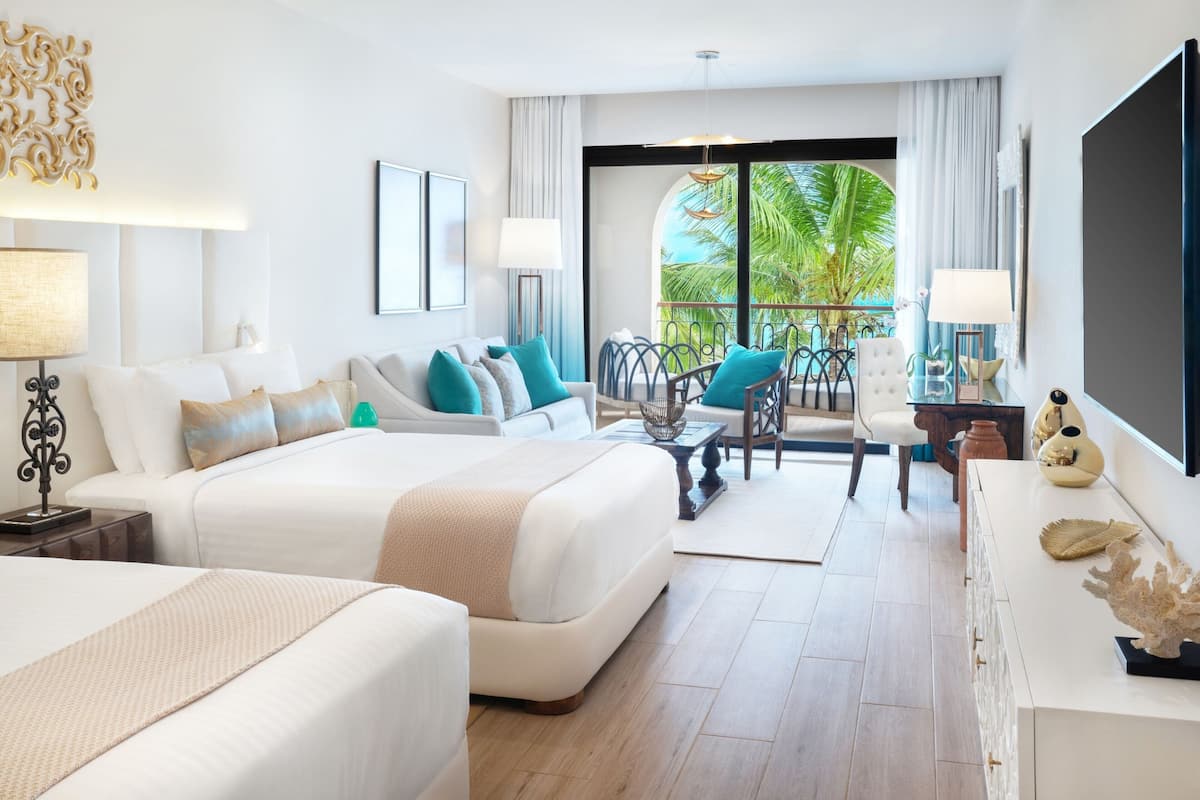A hotel room with a large bed, a sofa, a dining table, and a balcony with a view of palm trees.