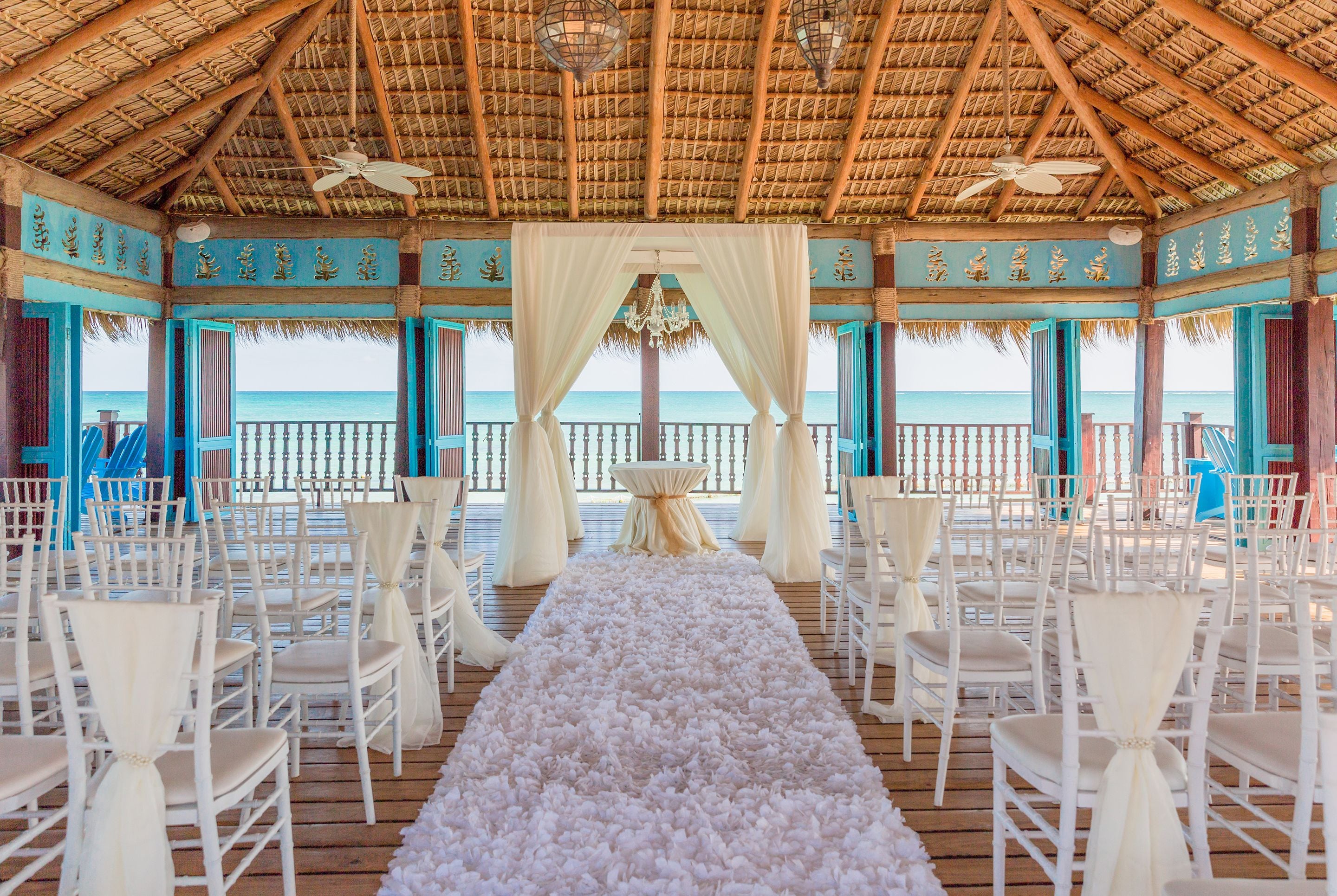 An outdoor wedding venue with a wooden deck, white chairs, a floral aisle, and a view of the ocean.