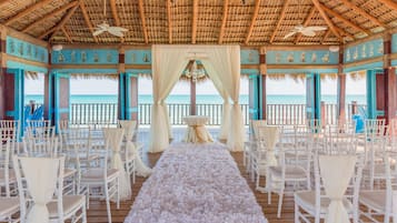 An outdoor wedding venue with a wooden deck, white chairs, a floral aisle, and a view of the ocean.
