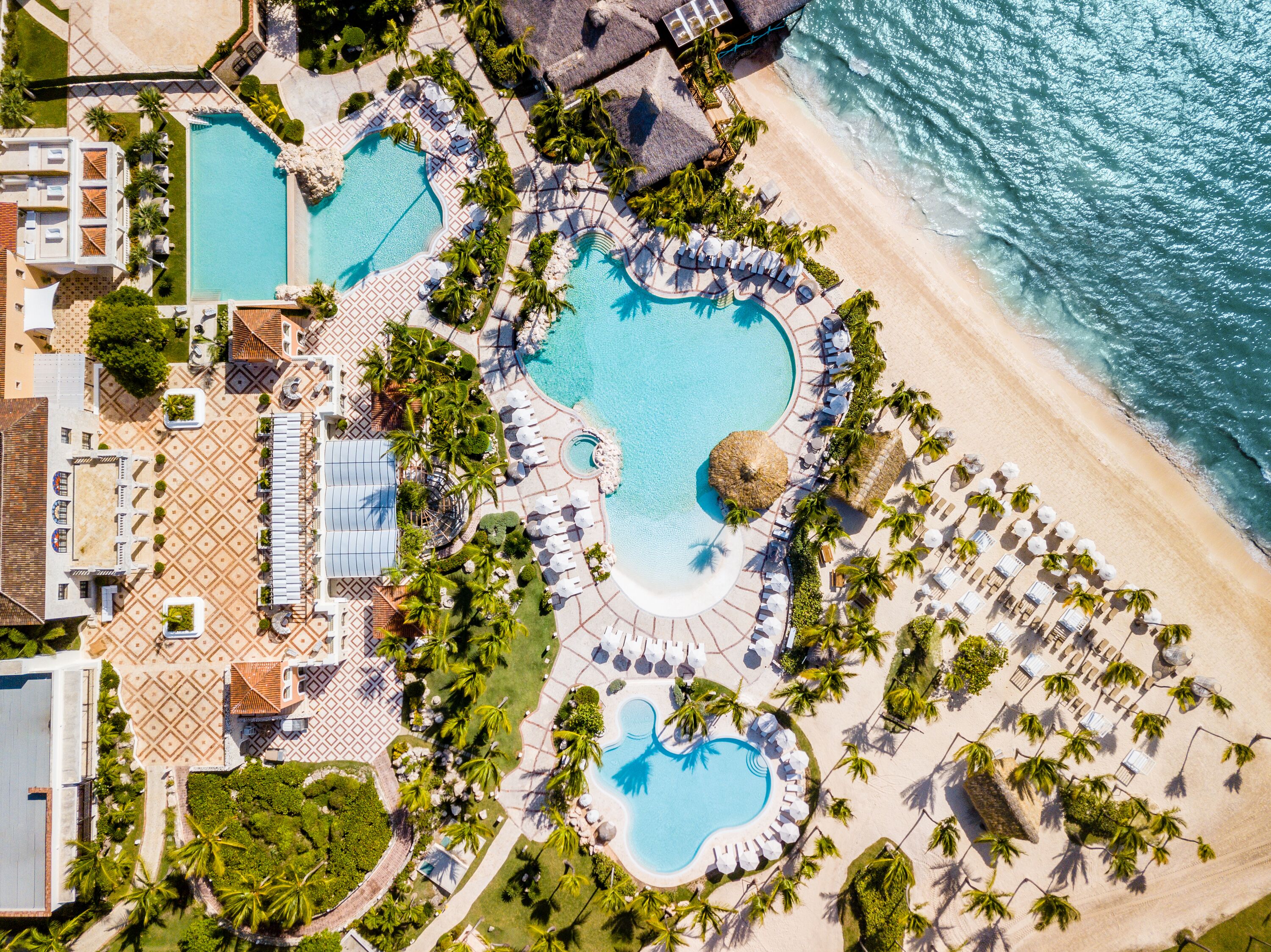 An aerial view of a resort with multiple swimming pools, beachside lounge areas, and palm trees.