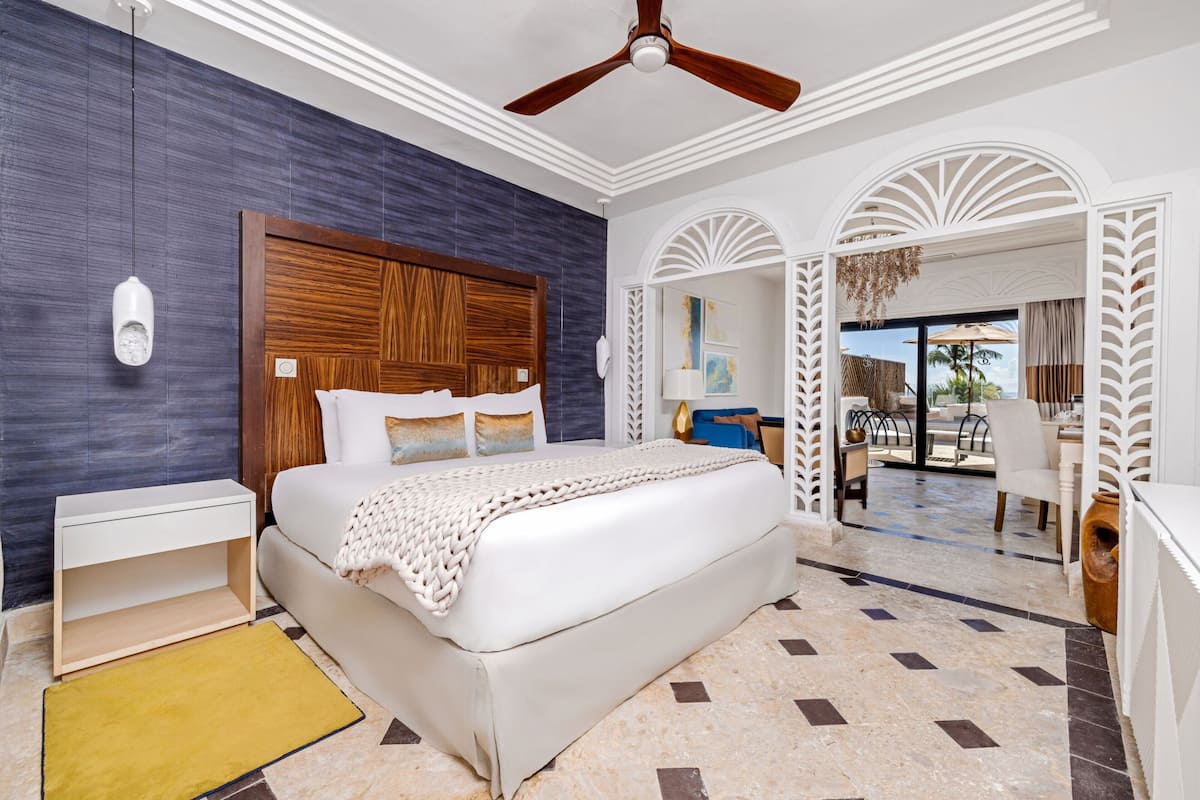 A modern hotel room with a large bed, a ceiling fan, a nightstand, and a view of the outdoor seating area.