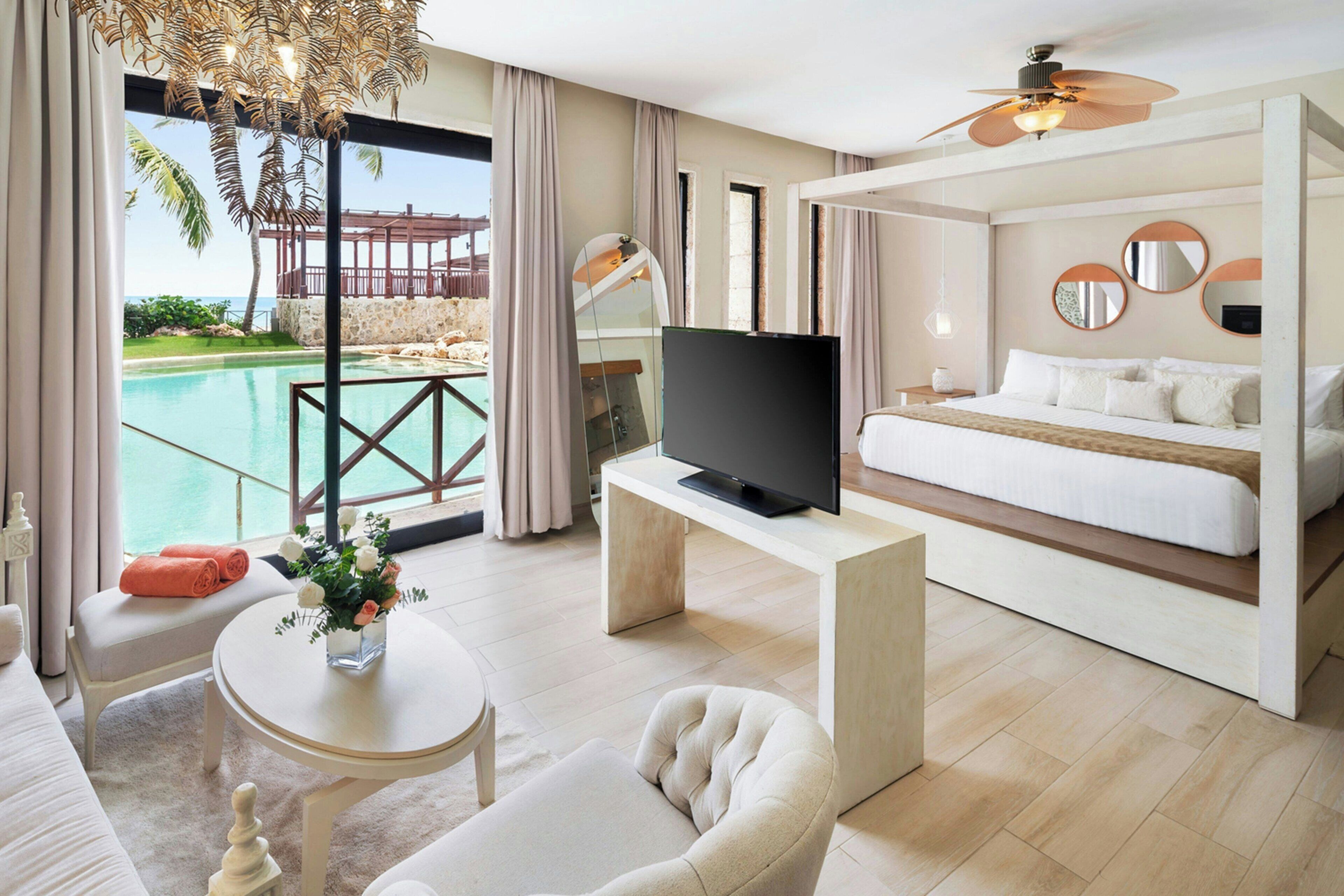 A modern hotel room with a large bed, a television, a seating area with a table, and a view of a pool and beach.