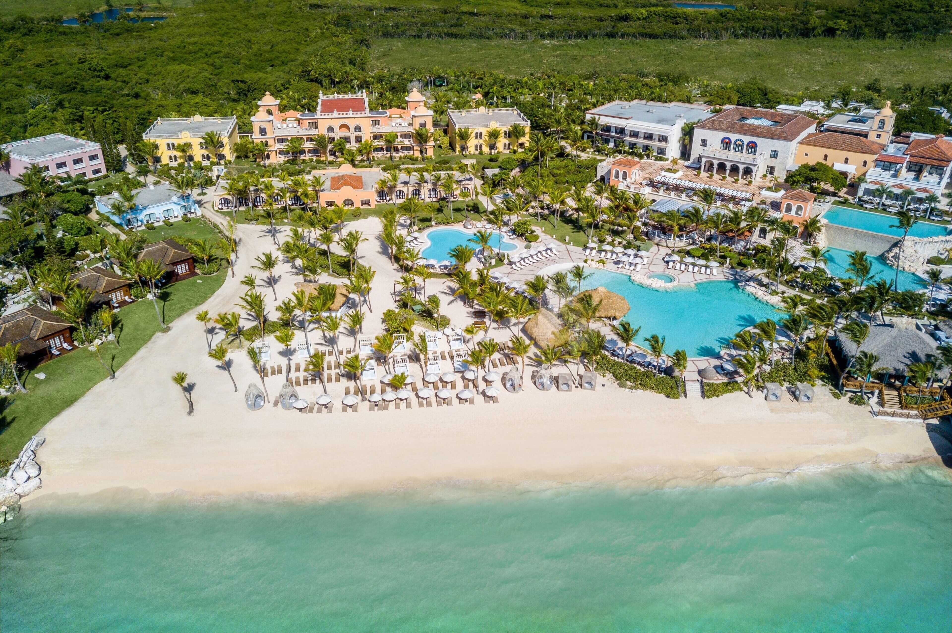 An aerial view of a resort with multiple buildings, a large central pool, and a beach area with sun loungers and palm trees.