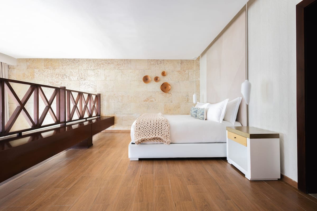 A modern bedroom with a wooden floor, a white bed with a beige throw, a nightstand, and a wooden railing.