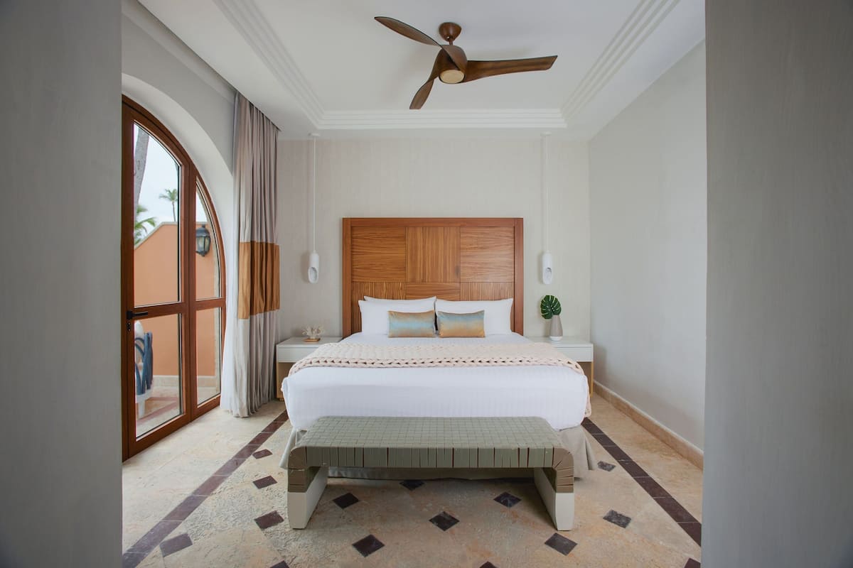 A hotel room with a large bed, a wooden headboard, a bench at the foot of the bed, a ceiling fan, and a view through a glass door.