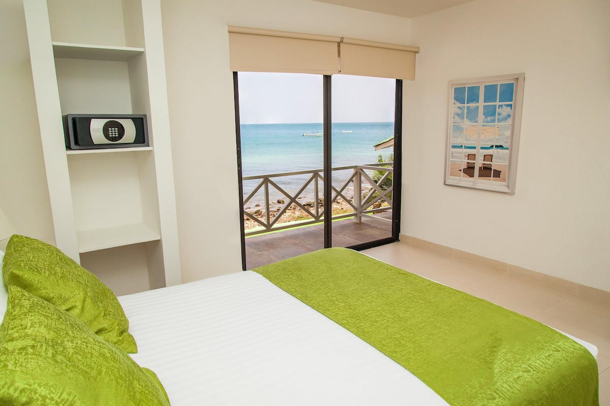 Un dormitorio con una cama, un almohadón verde, un almohadón blanco, un estante con un dispositivo, una ventana con vista a la playa y un cuadro en la pared.