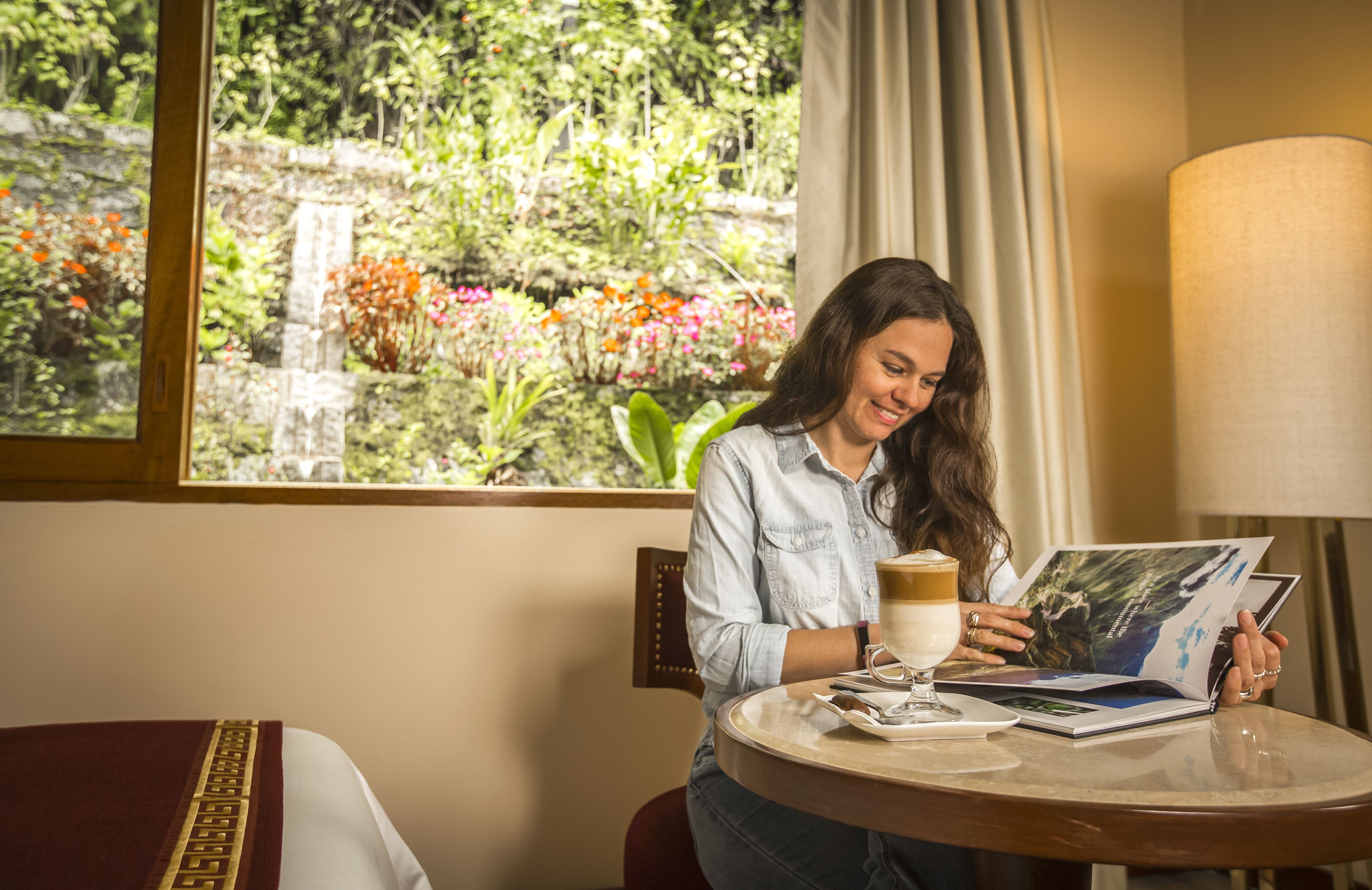Uma mulher sentada à mesa redonda com um livro e uma xícara de café, vista para a janela com vegetação e flores.