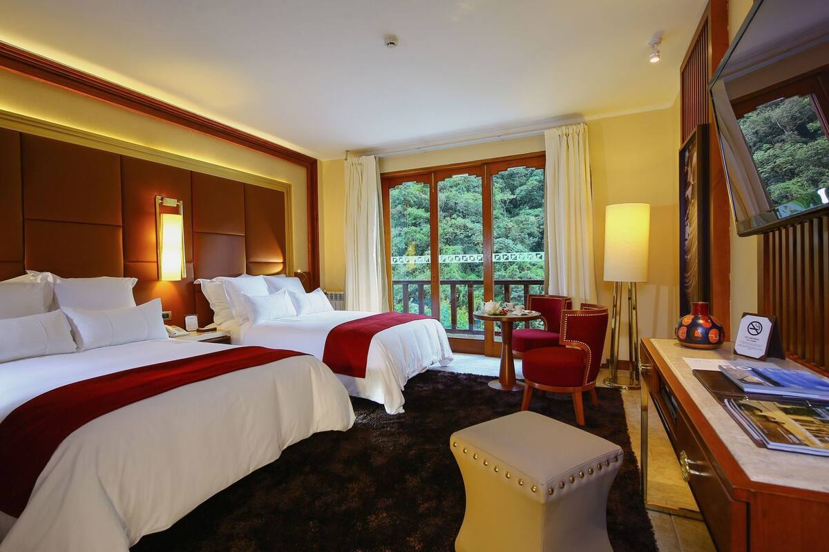 A hotel room with two beds, a desk, a chair, a TV, and a balcony with a view of trees.