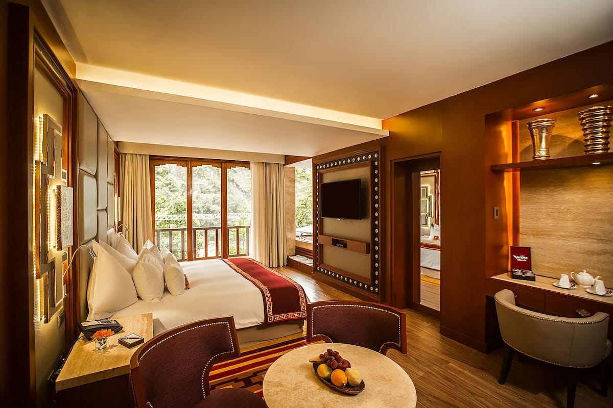 A hotel room with a bed, a TV, a dining area with a table and chairs, and a view of greenery outside.