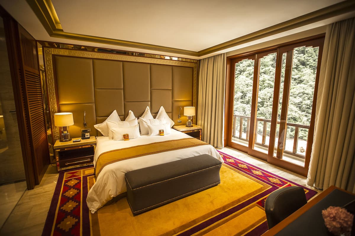 A hotel room with a large bed, a bench, a desk, and a view of trees through a large window.
