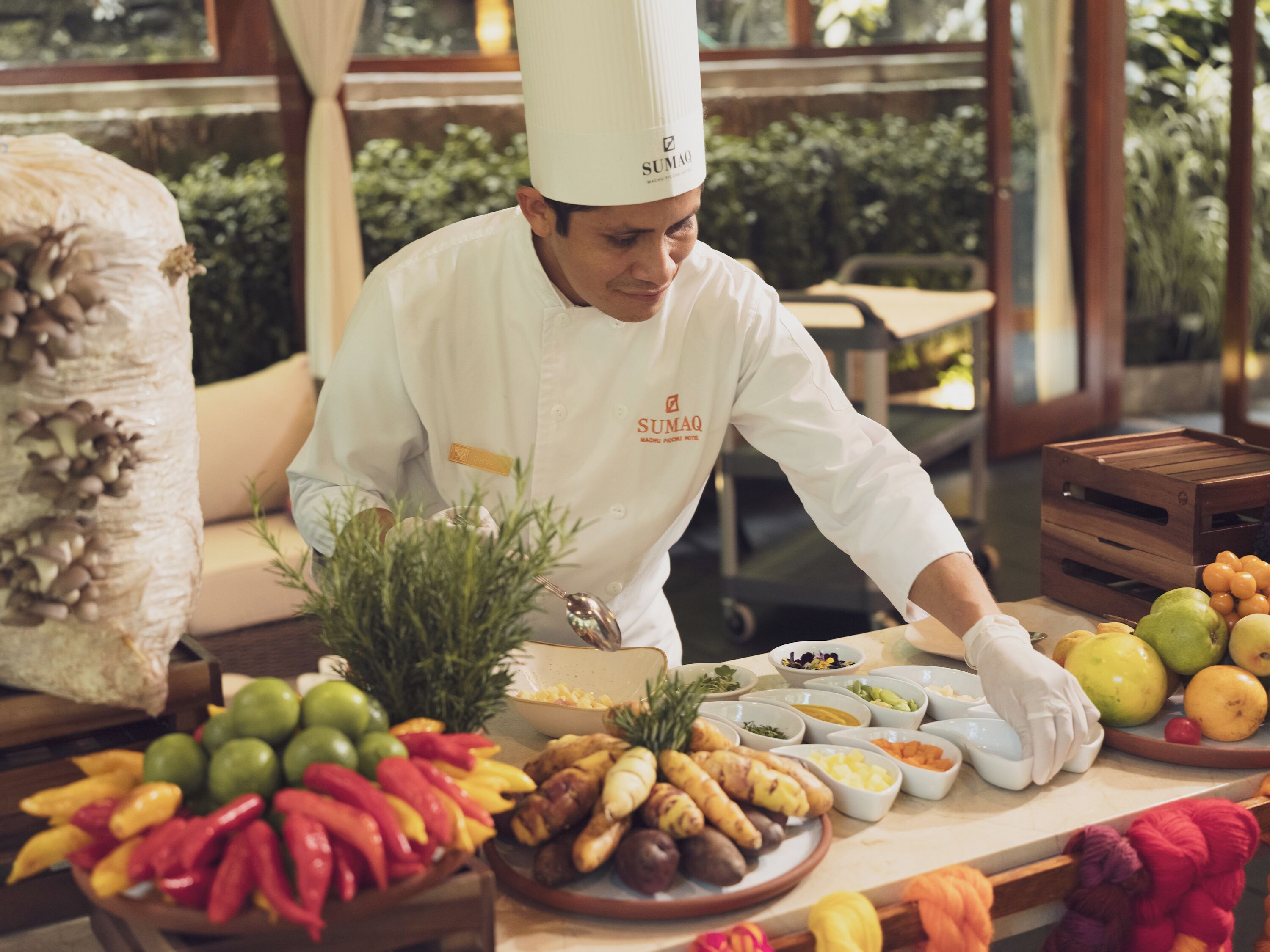 Um chef com uniforme branco e chapéu de chef preparando comida em uma mesa com diversas frutas e legumes.