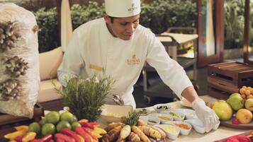 Um chef com uniforme branco e chapéu de chef preparando comida em uma mesa com diversas frutas e legumes.