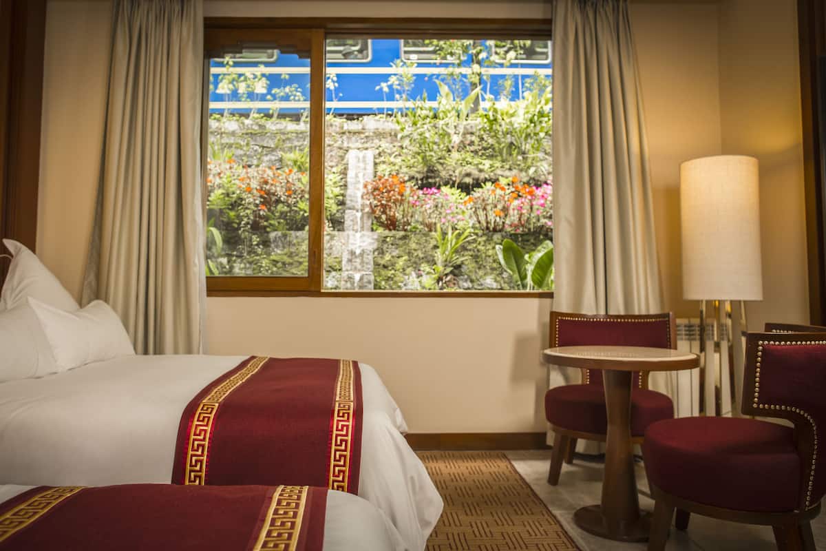 A hotel room with a bed, a small table, two chairs, a lamp, and a window with a view of a blue train and greenery.