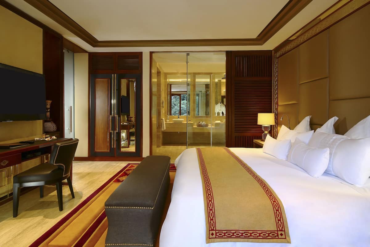 A hotel room with a large bed, a desk, a chair, a bench, a TV, and a mirror.