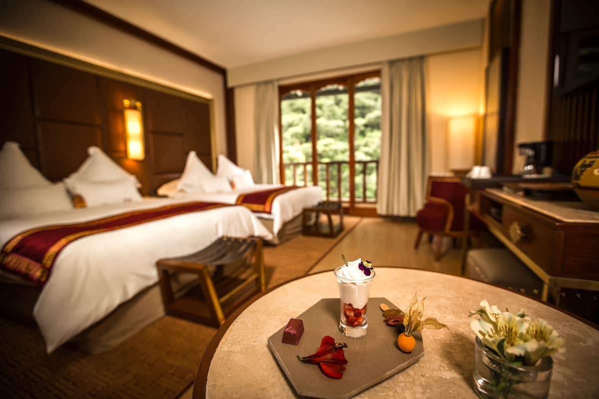 A hotel room with two beds, a dining table with a dessert, and a view of greenery outside.