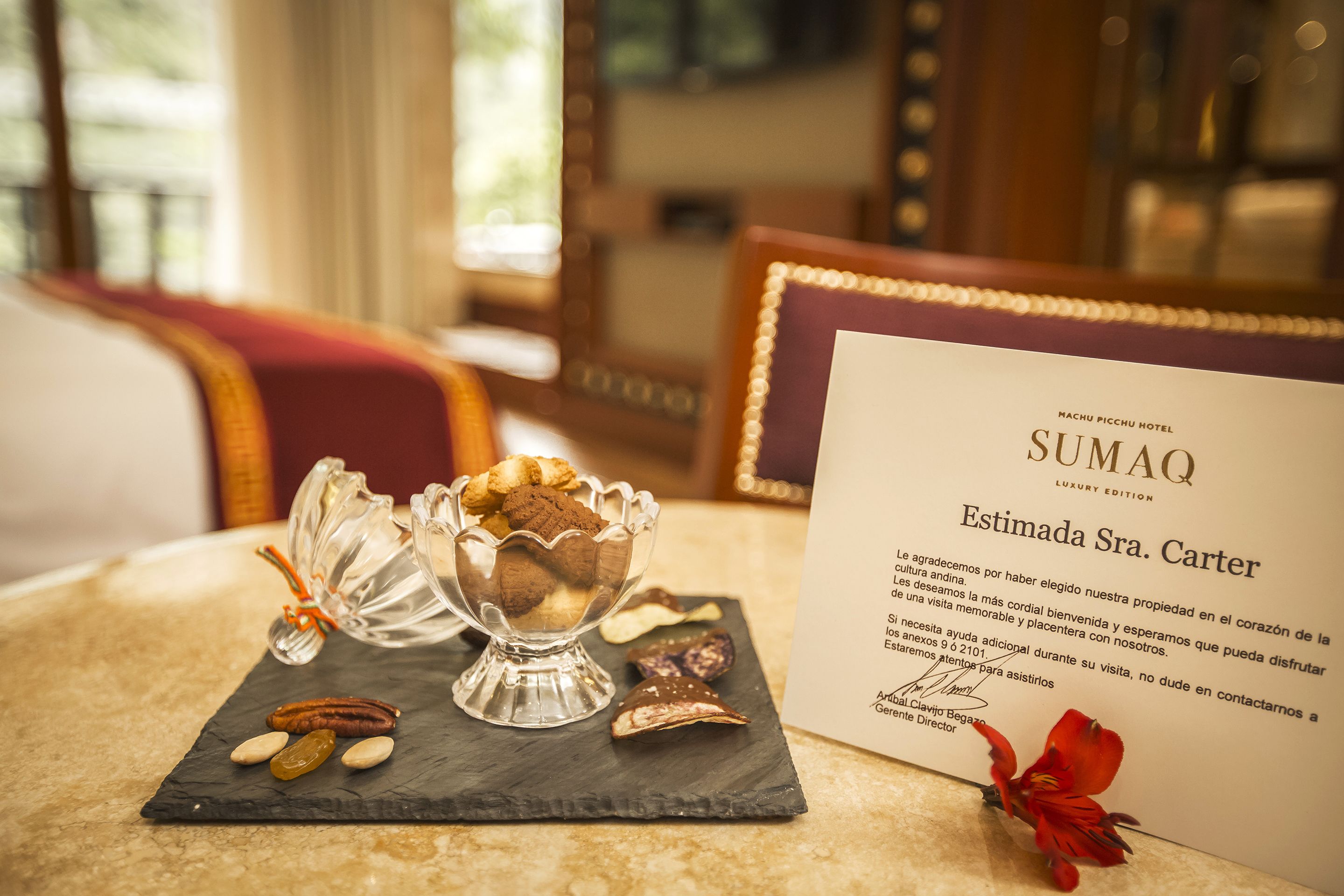 Una mesa de postres con galletas y frutos secos, un menú de Sumaq Luxury Edition y una flor roja sobre una mesa de mármol.