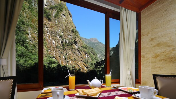 A breakfast table with a view of a mountainous landscape through large windows, a teapot, cups, and a tray of food. - Sumaq Machu Picchu Hotel (Machu Picchu)