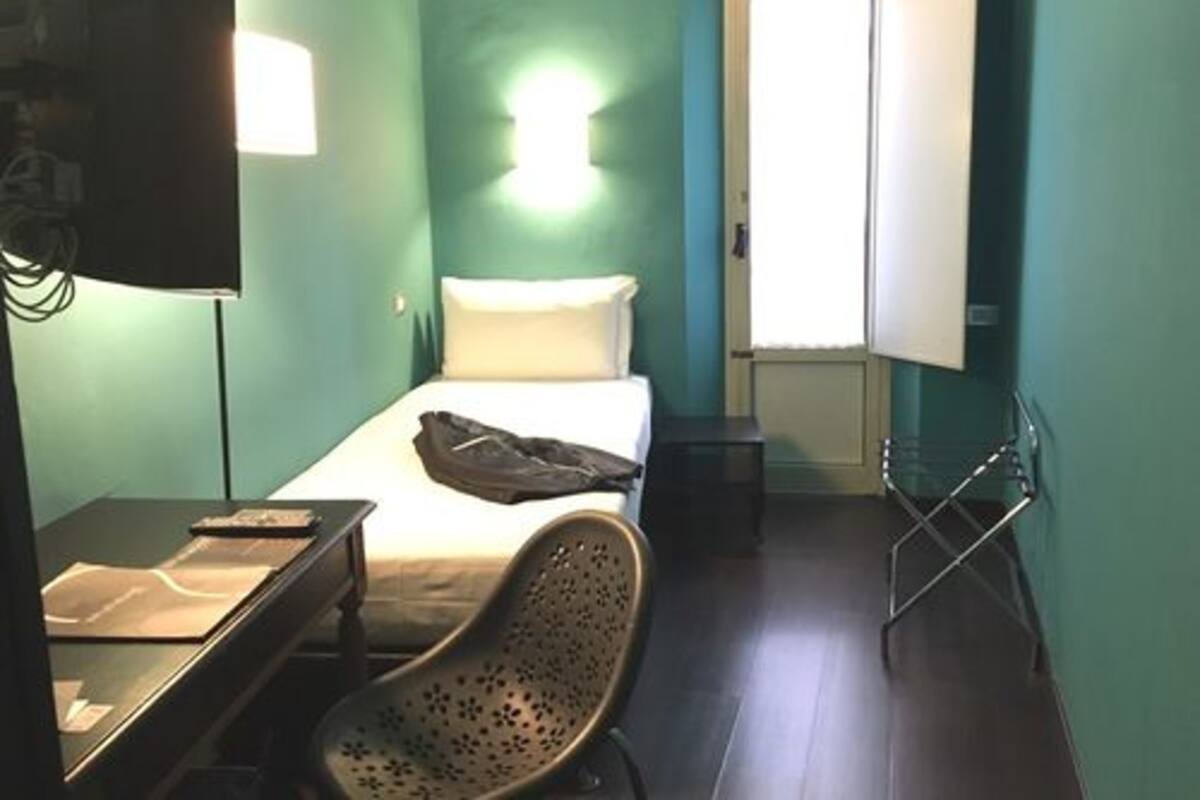 A small, single-bed room with a desk and chair, a window with blinds, and a wall-mounted TV.