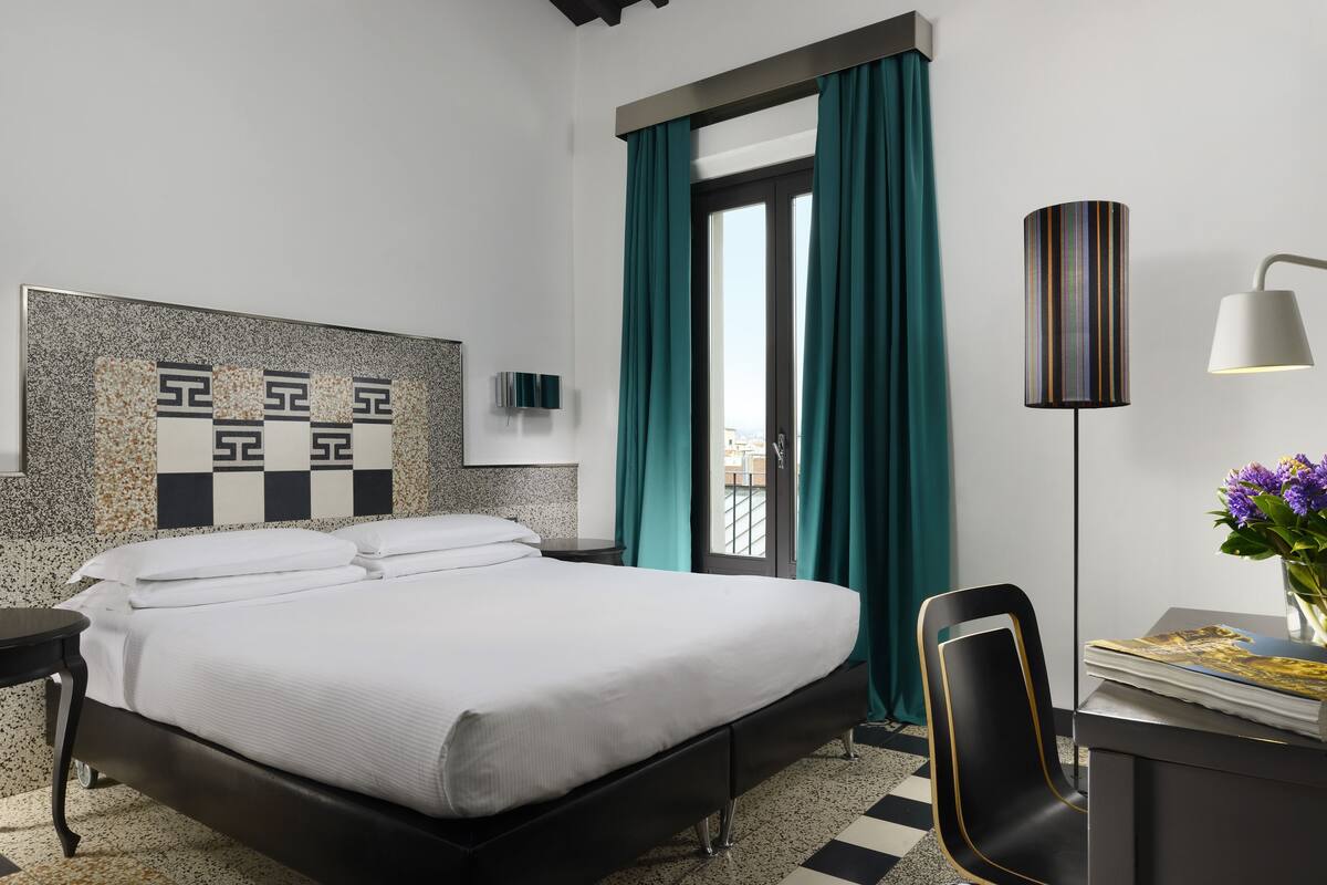 A modern hotel room with a large bed, a desk with a lamp, and a view through a glass door.