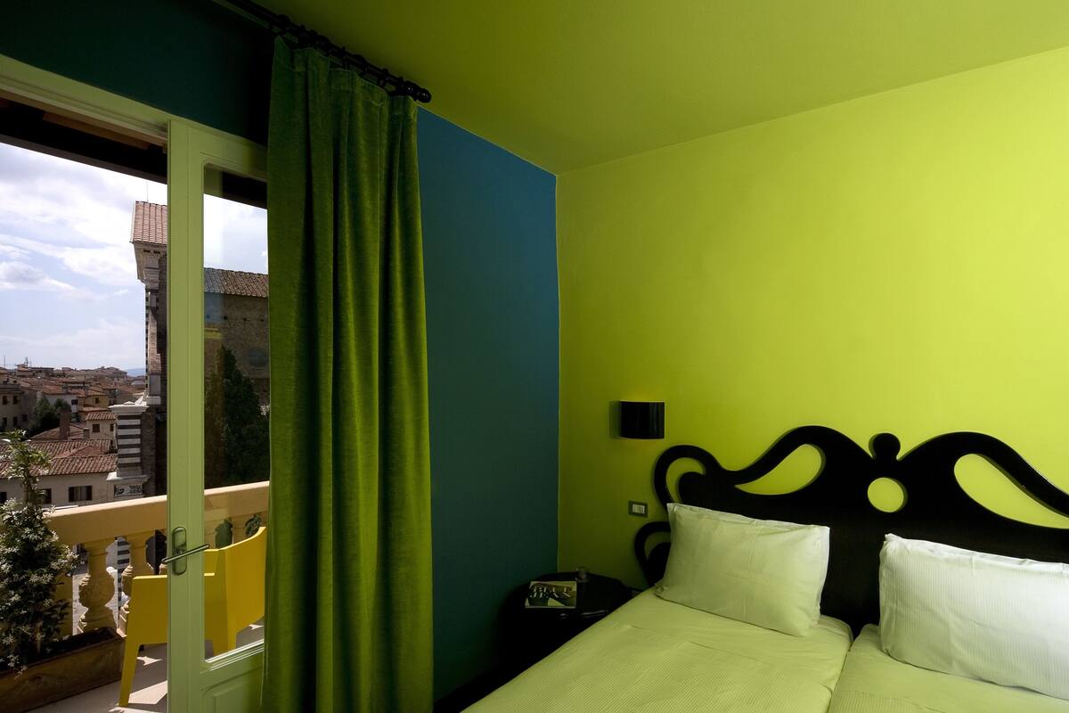 A room with a bed, a nightstand, and a view of a cityscape through a window with green curtains.