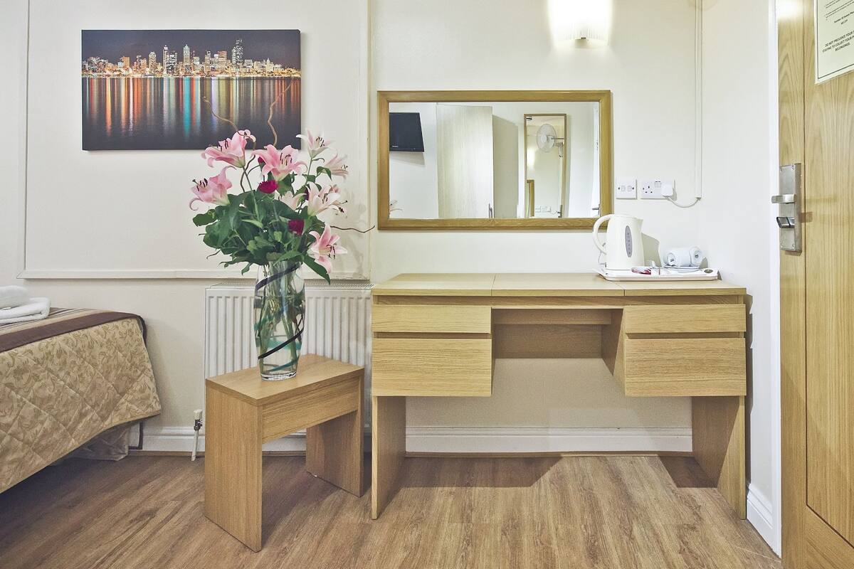 A hotel room with a wooden desk, a mirror, a vase of flowers, and a painting on the wall.