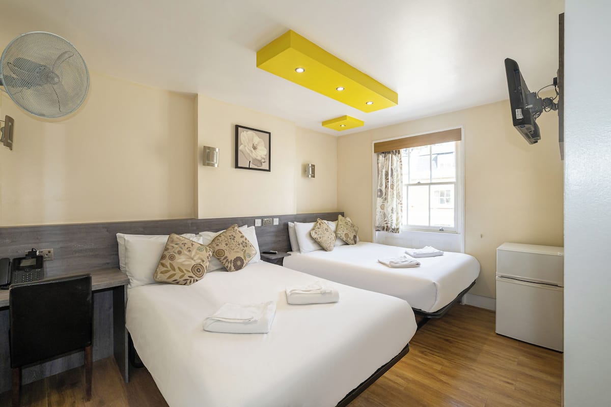 A hotel room with two beds, a desk, a chair, a small refrigerator, a ceiling fan, and a window with curtains.