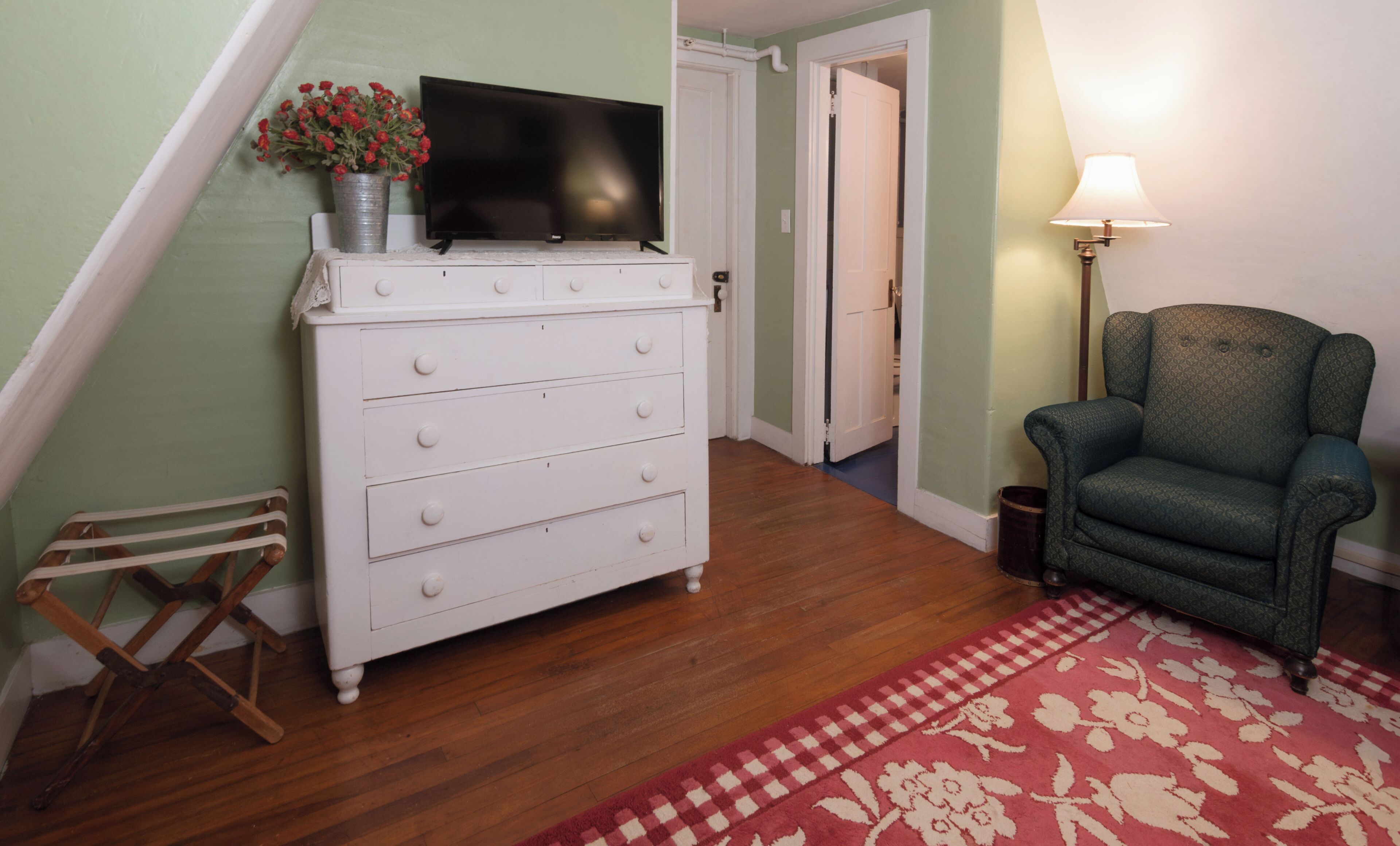 Una habitación con un tocador blanco, un televisor encima, un sillón verde y una alfombra roja estampada.