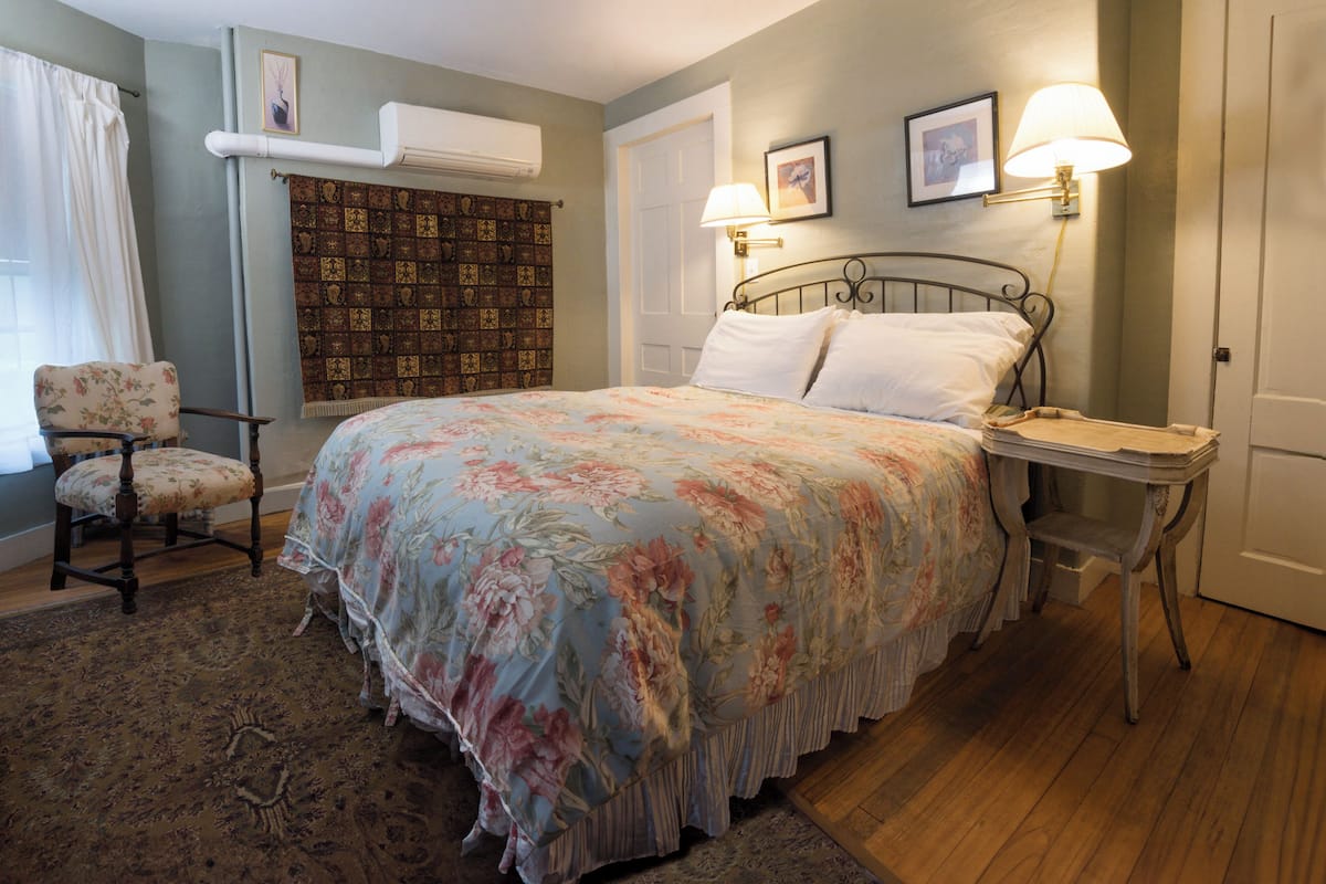 A bedroom with a floral bedspread, a chair, a bedside table, and a wall-mounted lamp.